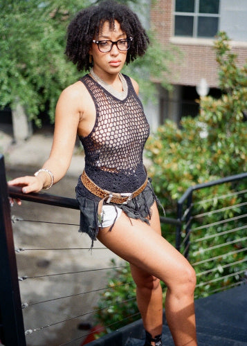 Woman wearing a fishnet top and denim shorts leaning against a railing outdoors.